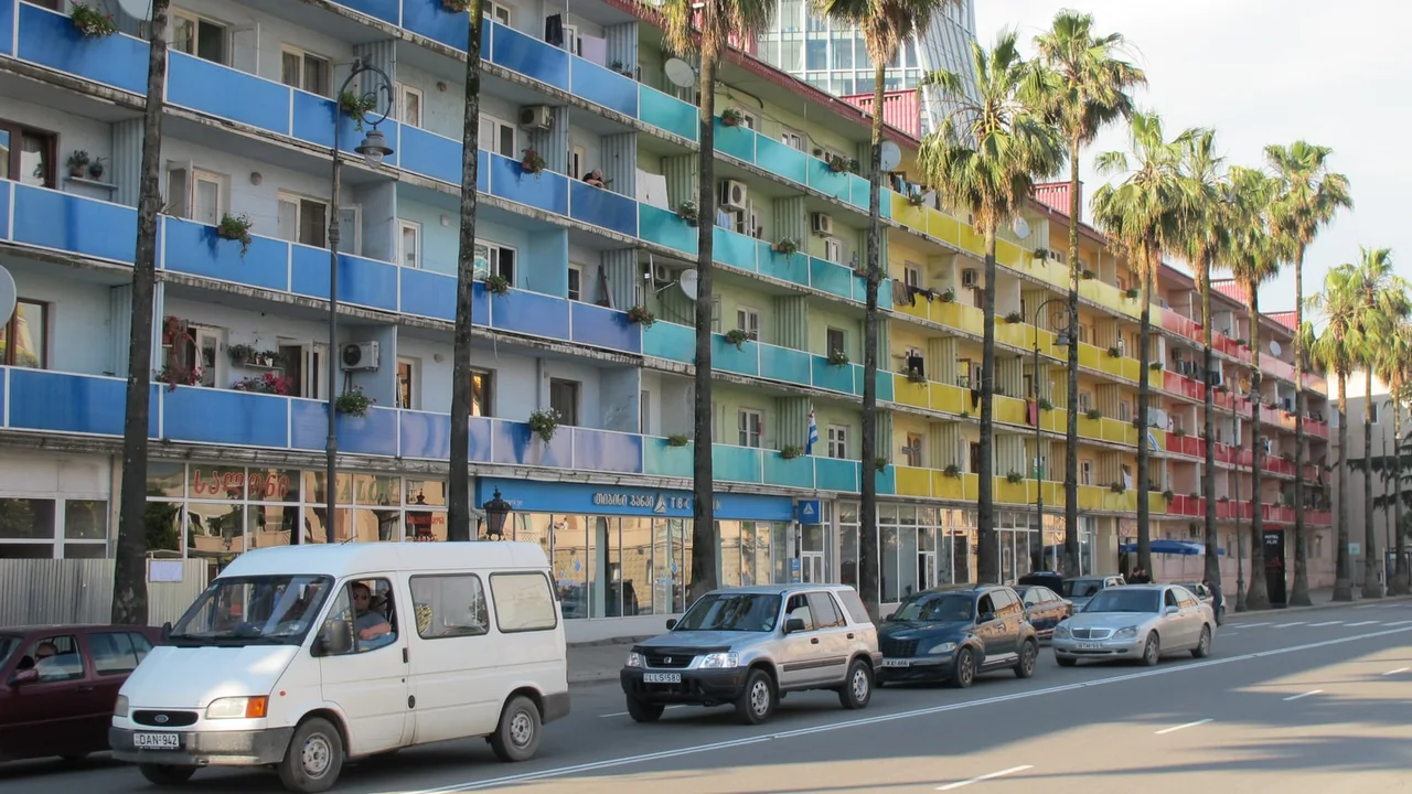 A white minibus and cars driving along Rustaveli Avenue in Batumi past an apartment block with multicoloured balconies and a row of palms