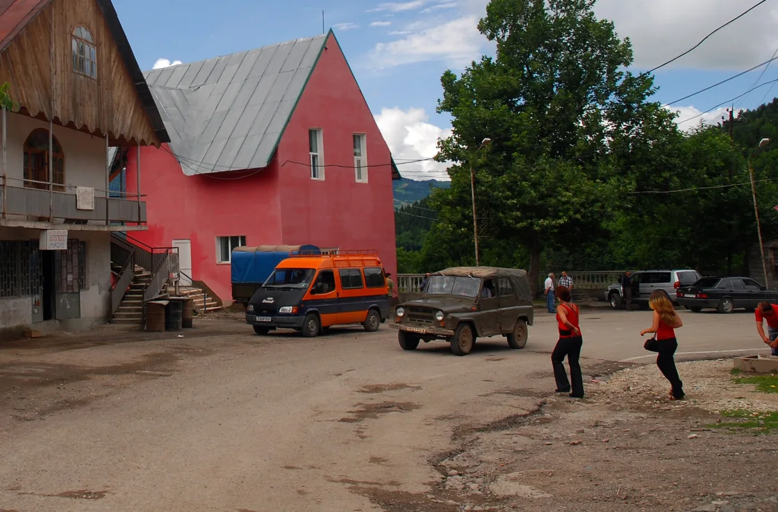 An orange minibus and a canvas-topped off-road vehicle on unpaved ground by the road in Khulo, a pink house with a grey roof behind
