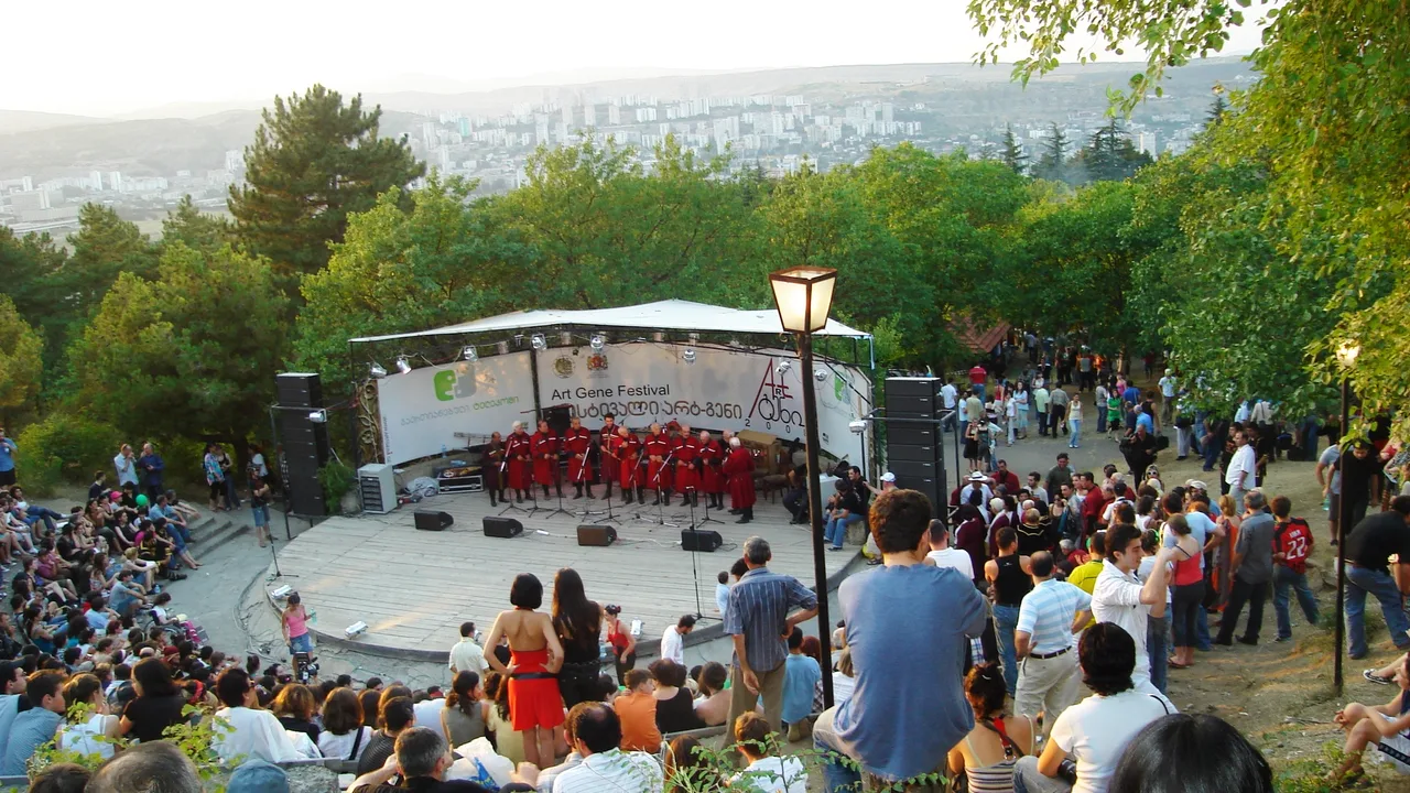 Folk ensemble in red chokhas on an open-air stage, crowd on the grassy slope and Tbilisi behind