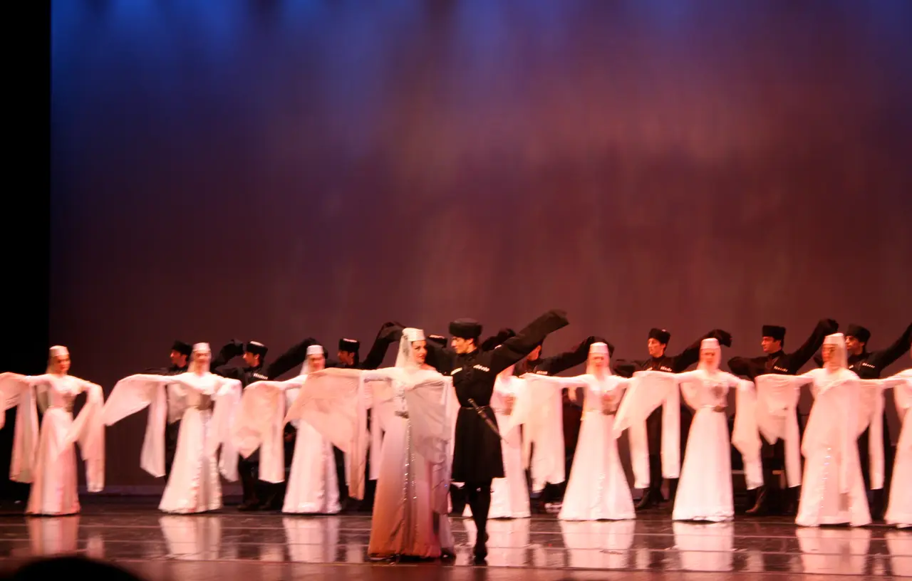 Row of dancers in white and black Georgian costumes with arms outstretched on a stage