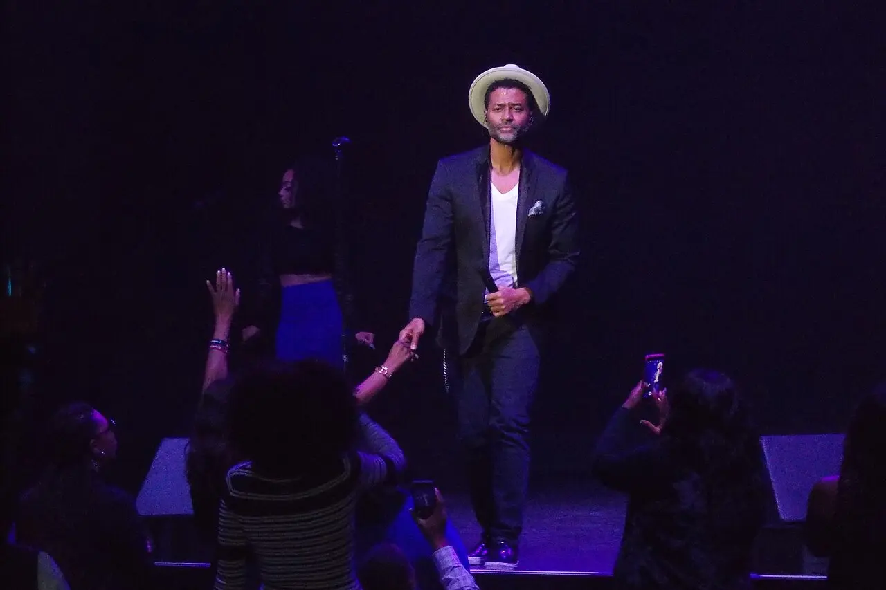 Singer in a pale hat and dark suit on a dim stage, phones raised in the front row