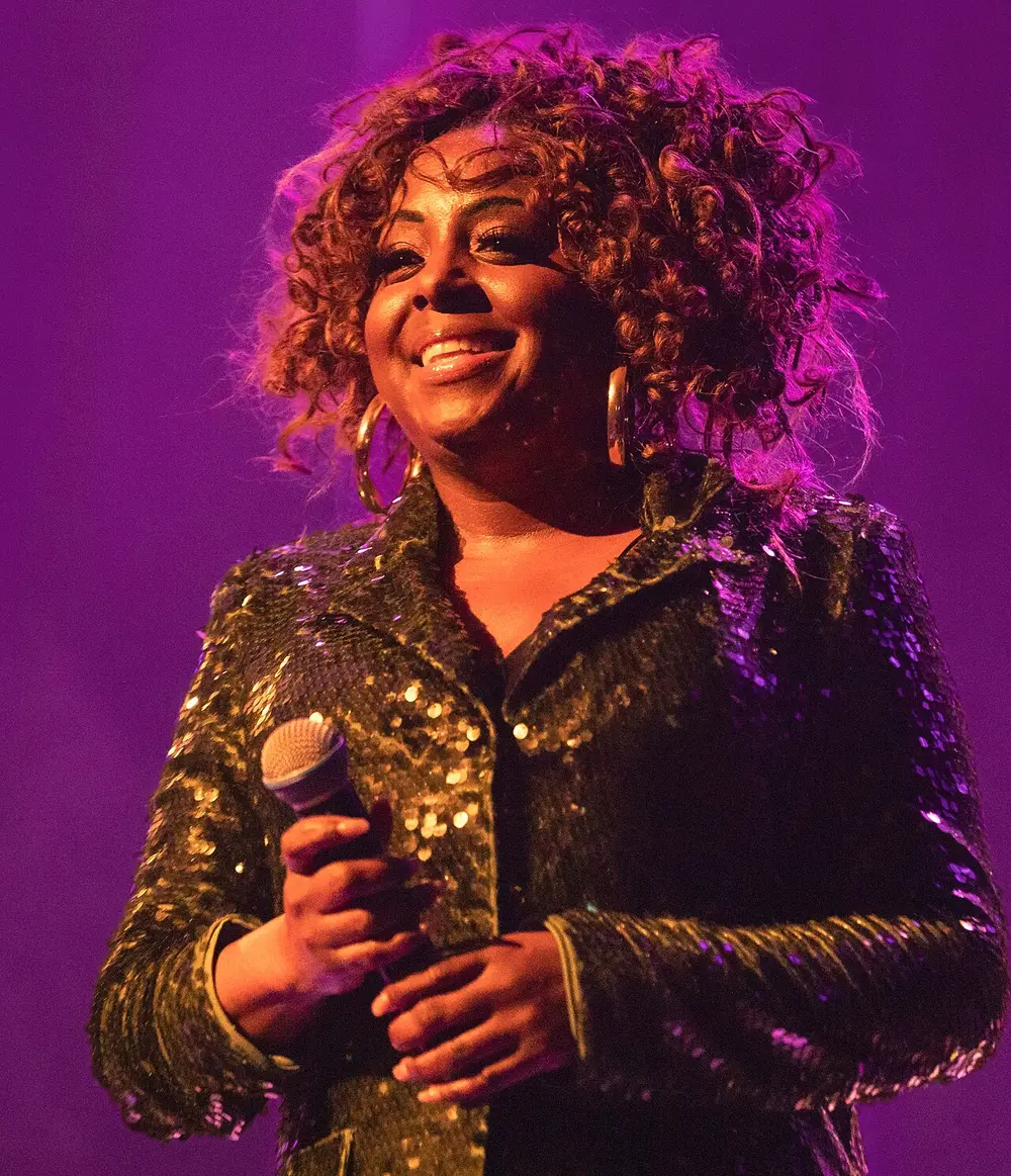 Singer in a sequinned jacket smiling with a microphone under purple stage light