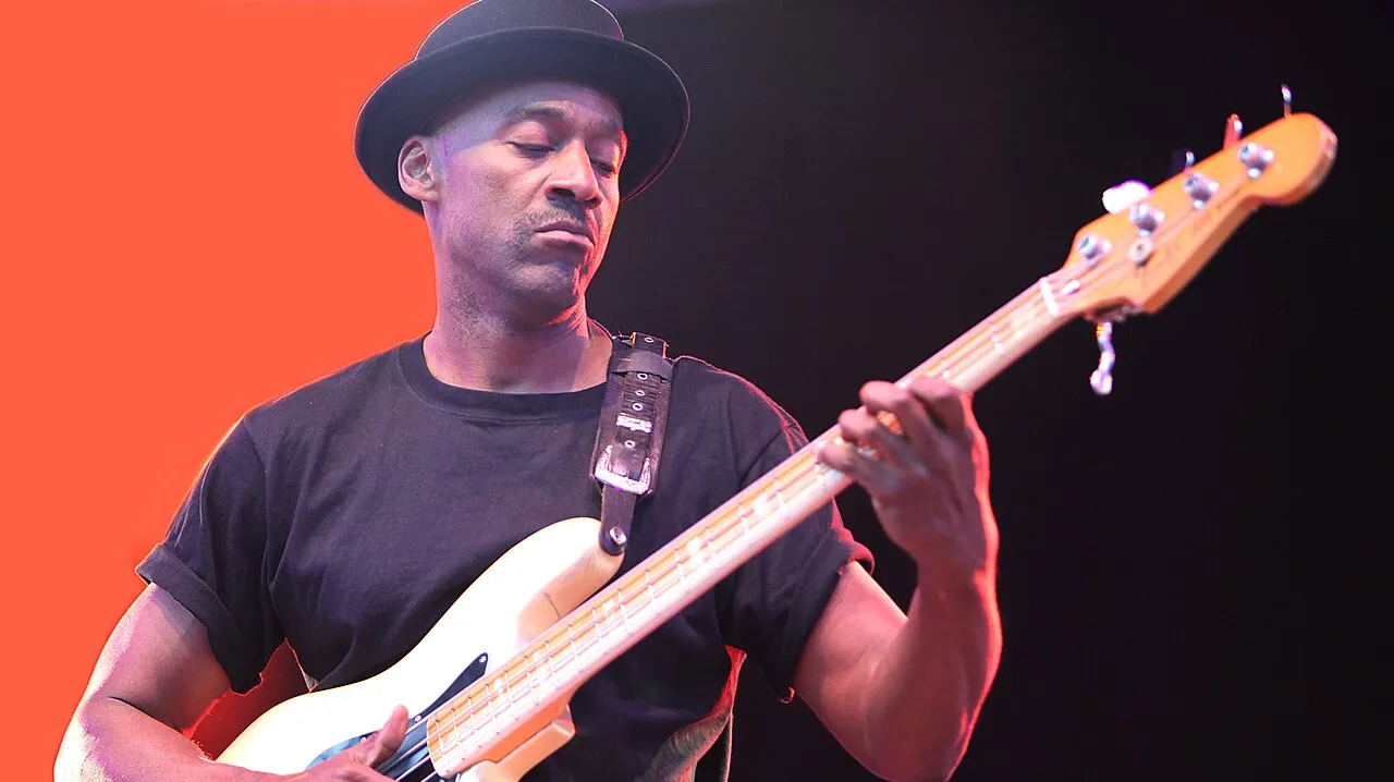 Bassist Marcus Miller in a bowler hat on a lit stage against an orange backdrop