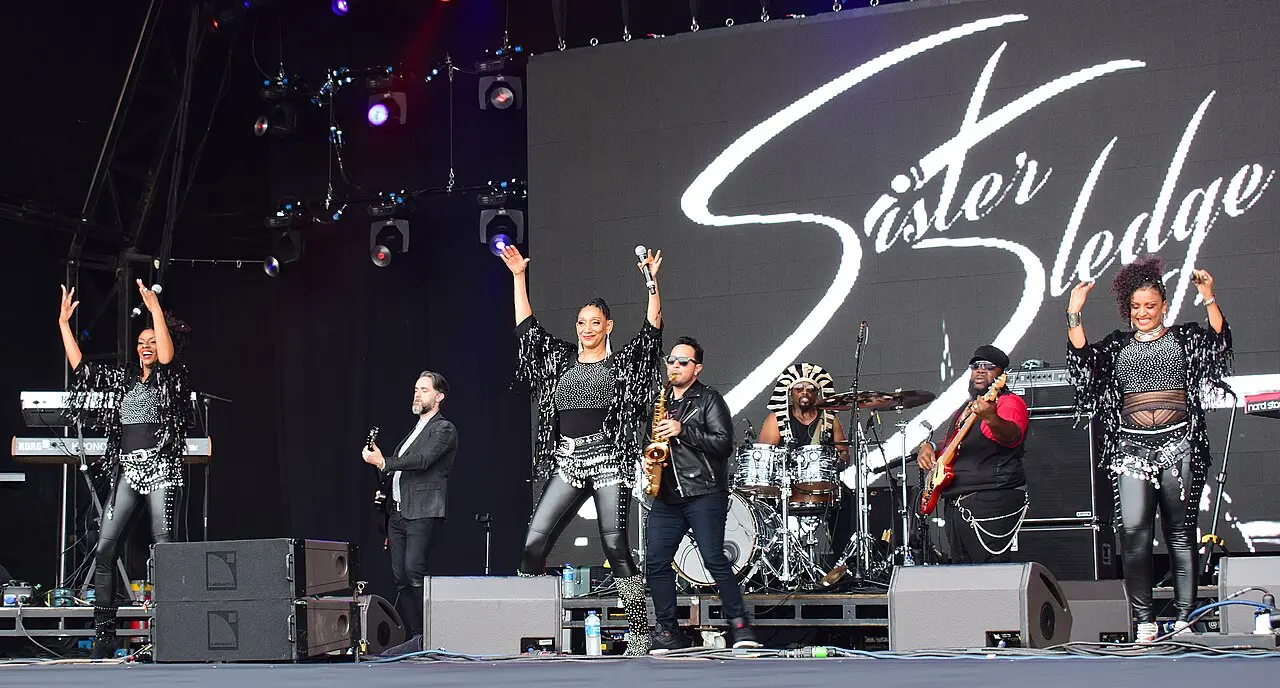 Band and three singers in sparkling outfits on a wide festival stage with a Sister Sledge backdrop