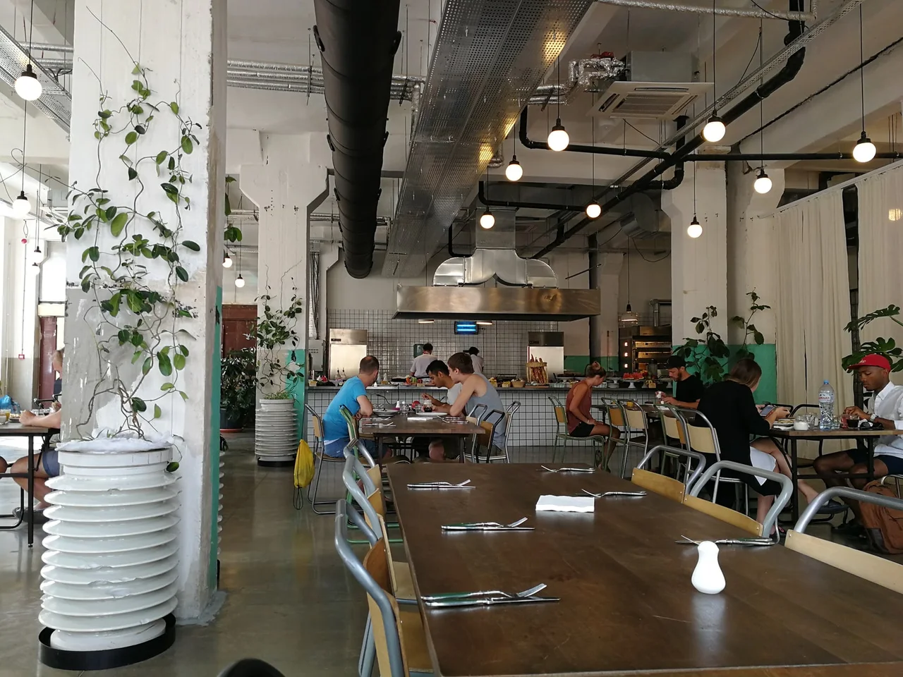 A cafe hall inside the former factory: white concrete columns, exposed ducts, pendant lamps and long wooden tables around an open kitchen