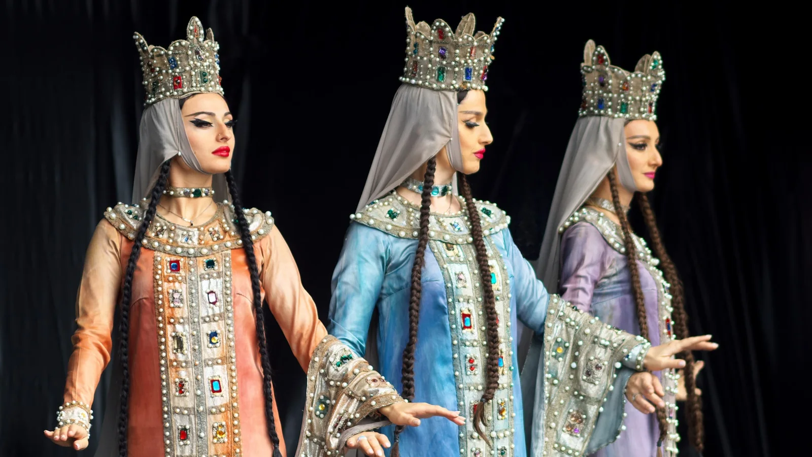 Three Sukhishvili dancers in jewelled crowns and long braids against a dark stage curtain