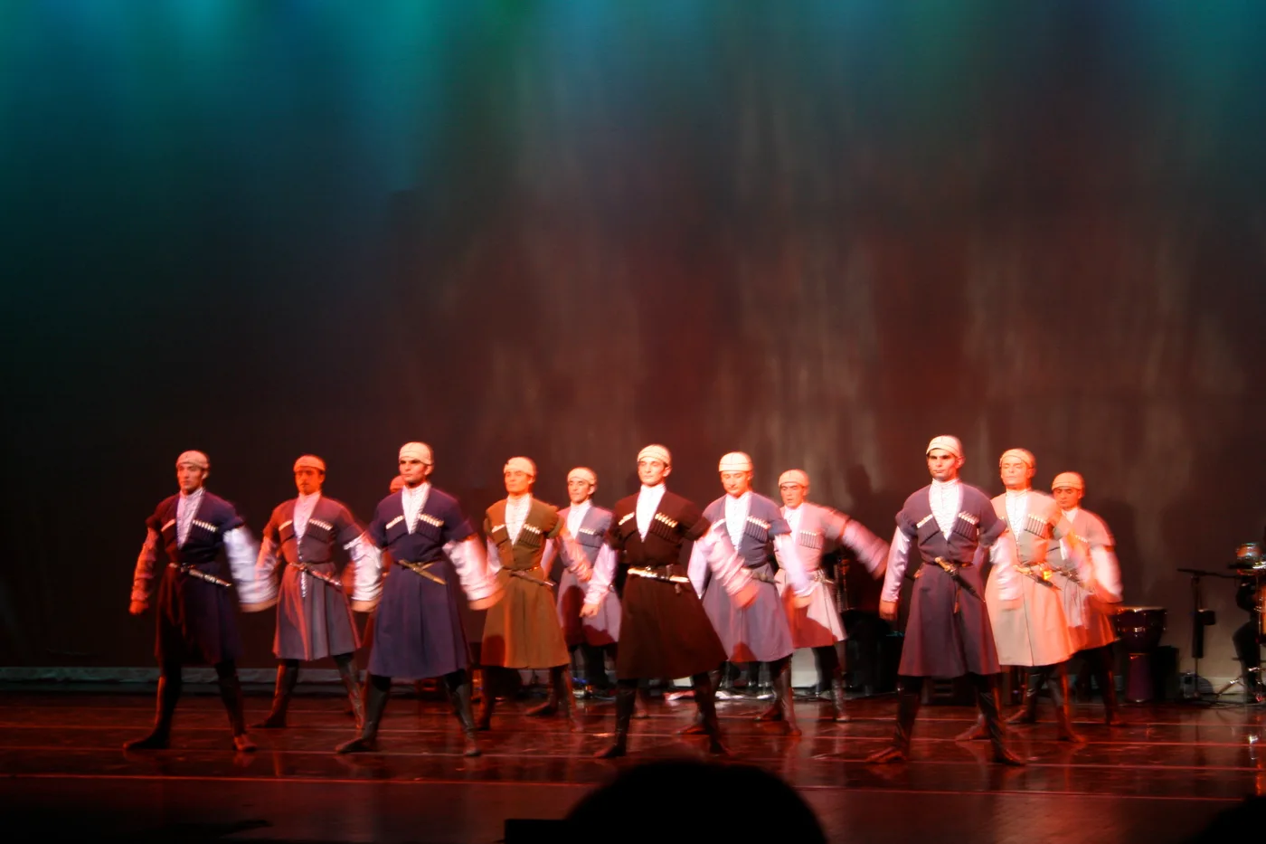 Davluri: a line of dancers in dark chokha coats and pale dresses on a lit stage