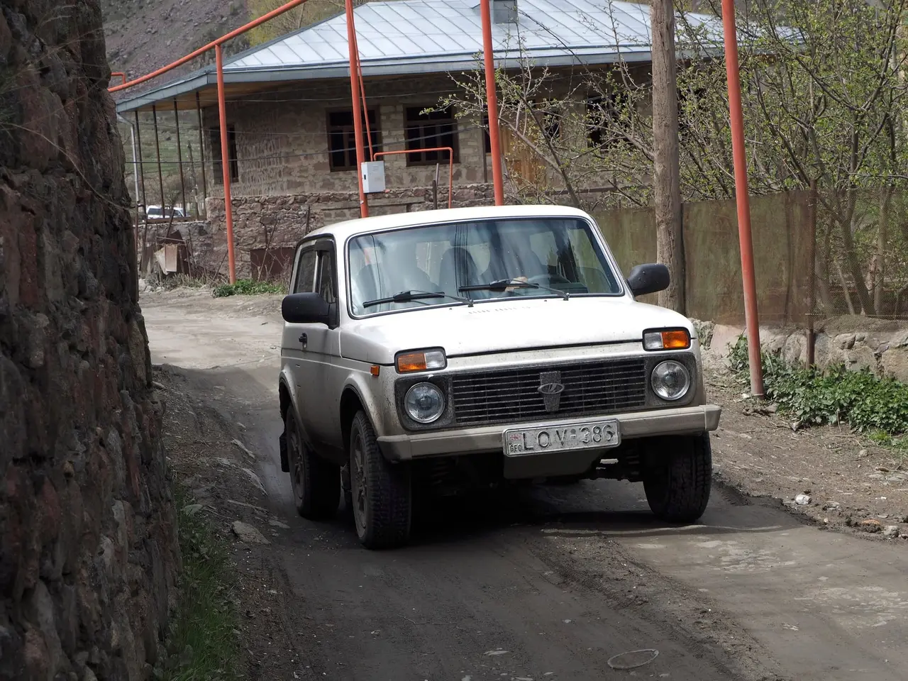 A white Lada Niva 4x4 with a Georgian plate on a dirt lane between a stone wall and a stone house