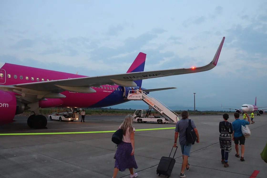 A Wizz Air plane parked at the steps on the apron of Kutaisi airport, with passengers carrying backpacks and a suitcase walking towards it