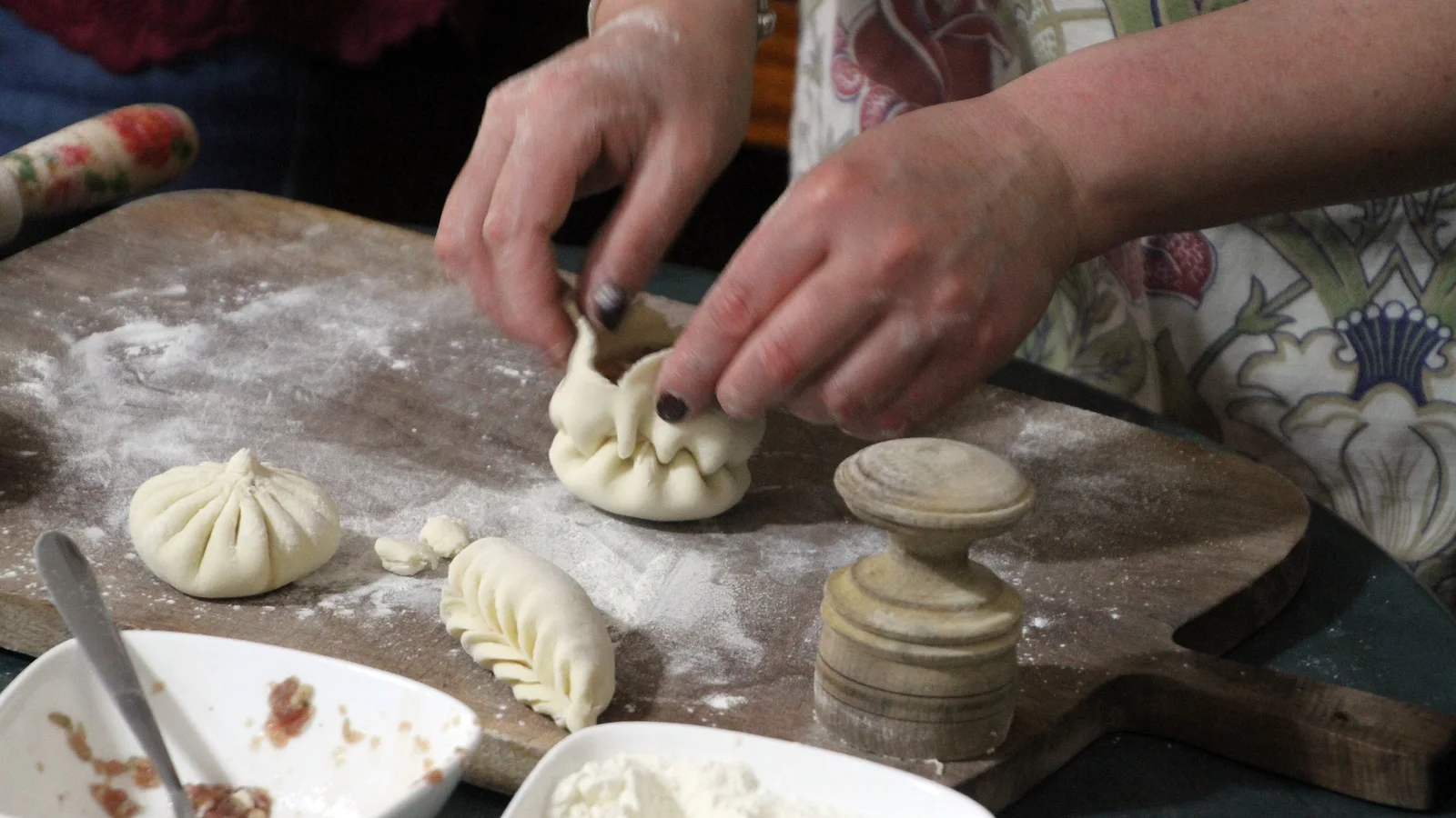 Hands pleating a khinkali on a floured wooden board, with a finished one and a bowl of filling beside it