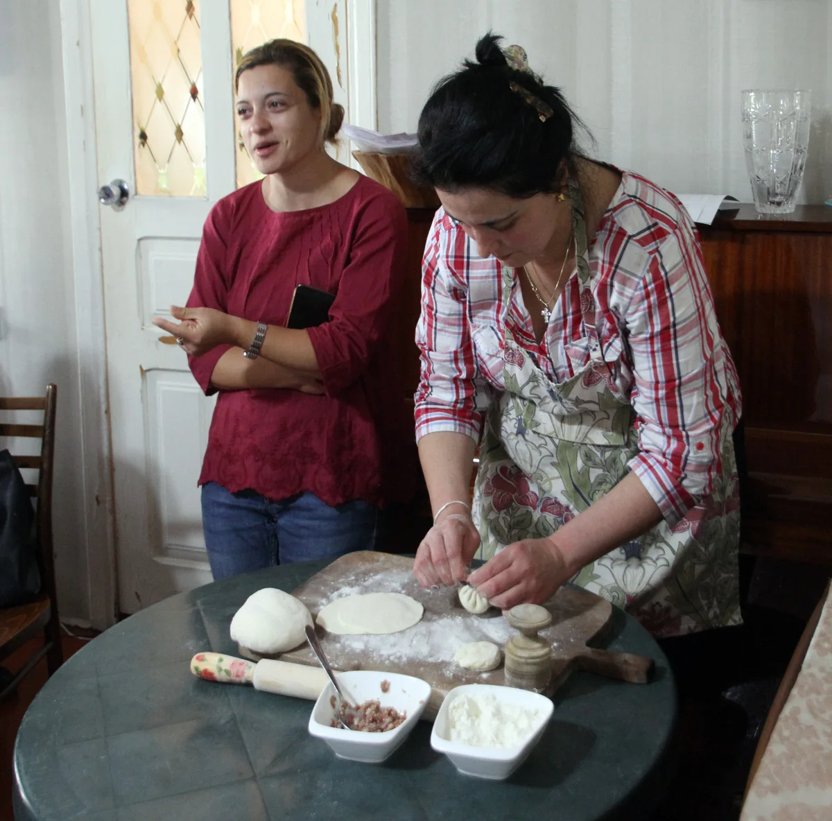 A woman shapes a khinkali at a round table while another woman stands by; dough, a board, a bowl of filling and a bowl of flour on the table