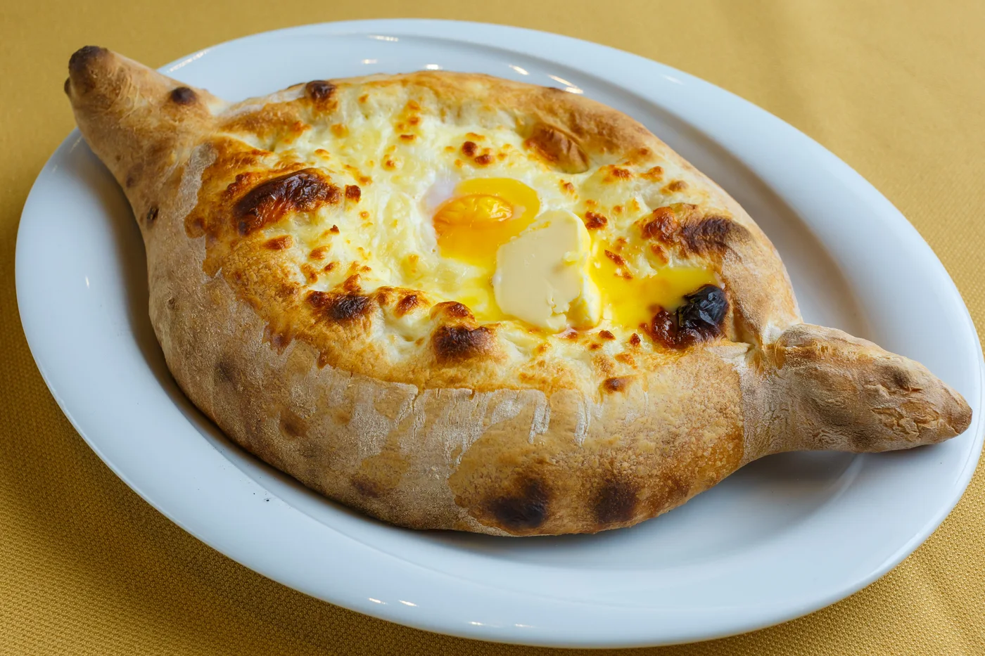 Adjarian khachapuri on a white oval plate: a boat of golden baked dough with melted cheese, an egg yolk and a pat of butter in the middle