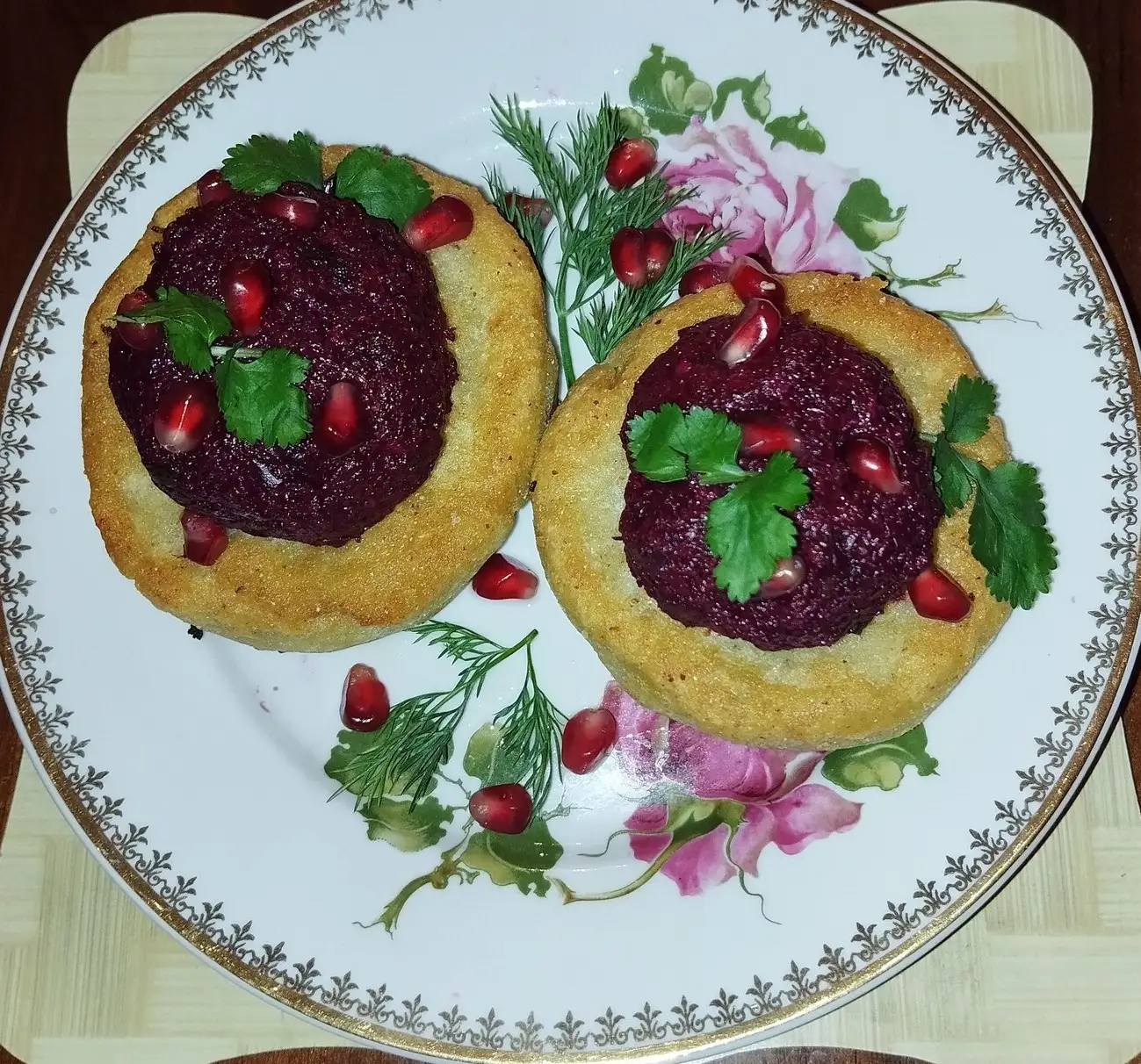 Two golden cornmeal cakes topped with dark red beetroot pkhali, pomegranate seeds and coriander on a flowered plate