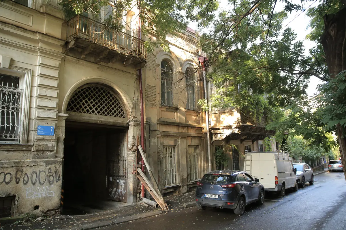 An arched courtyard gate with a lattice fanlight and a balcony above it on a late-19th-century house in Sololaki, with a carved wooden balcony among leaves next door and parked cars along the street