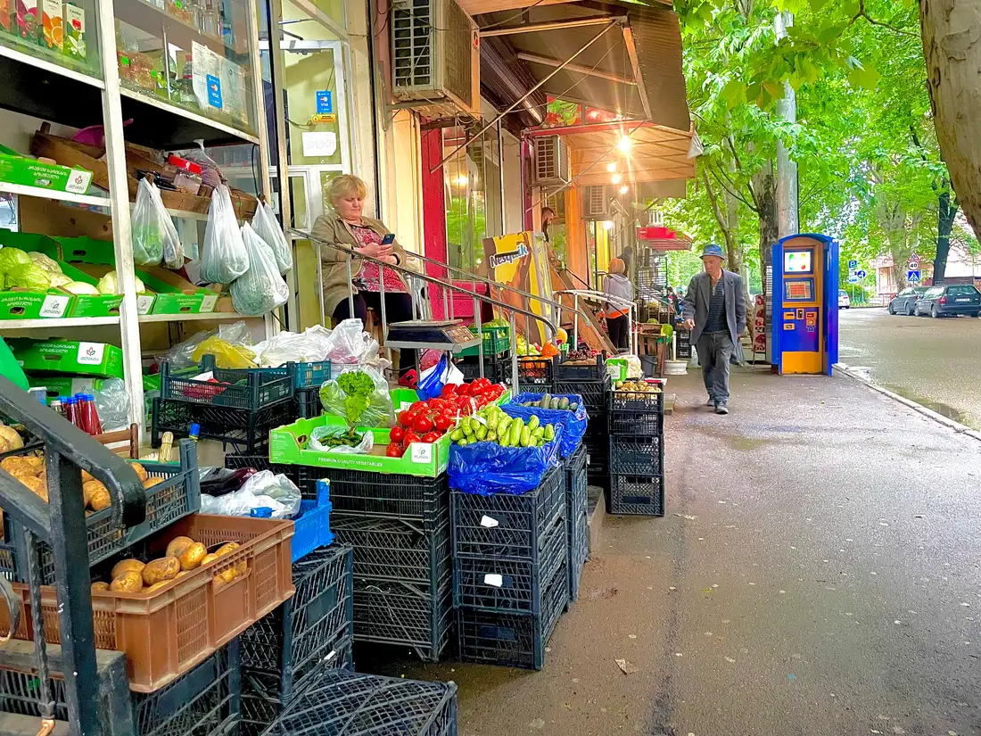 A greengrocer's stall on a Tbilisi pavement: crates of tomatoes, cucumbers, cabbage and potatoes, a seller sitting behind the scales and a man walking past