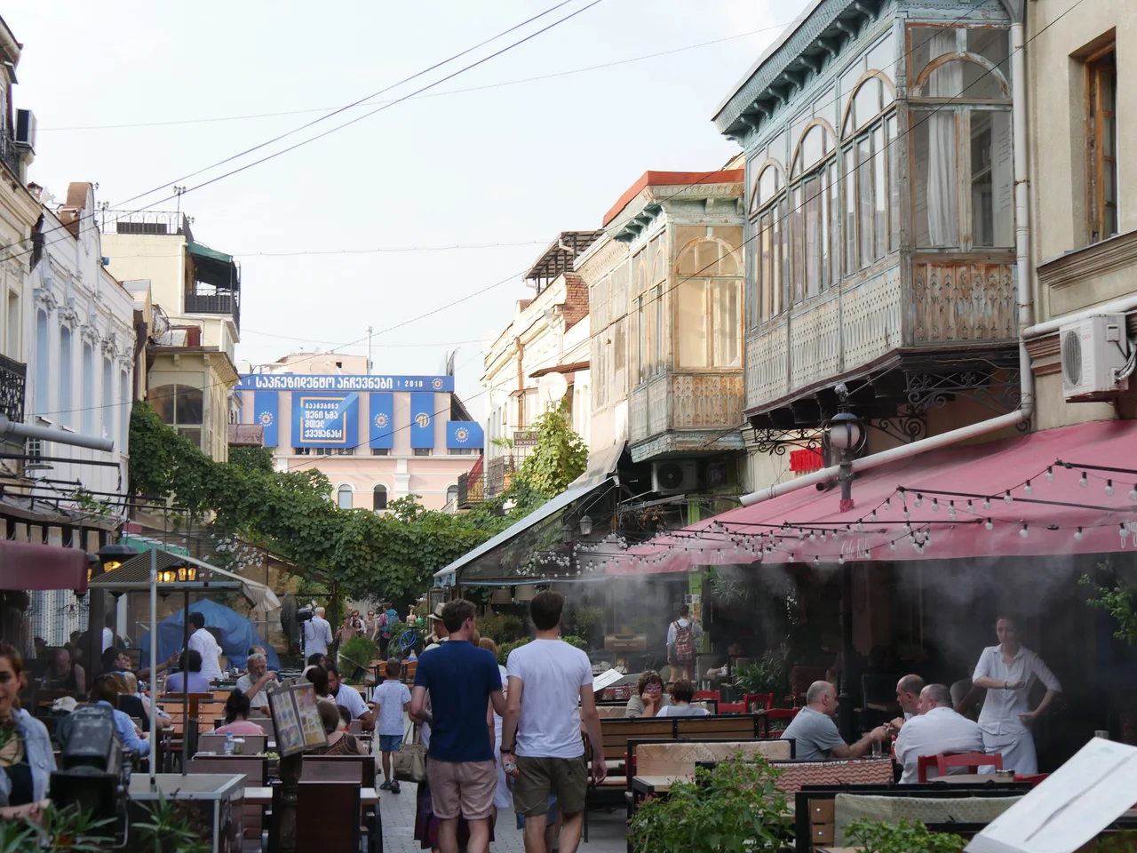 Cafe terraces with awnings and string lights along Erekle II Street in old Tbilisi, with people walking between the tables