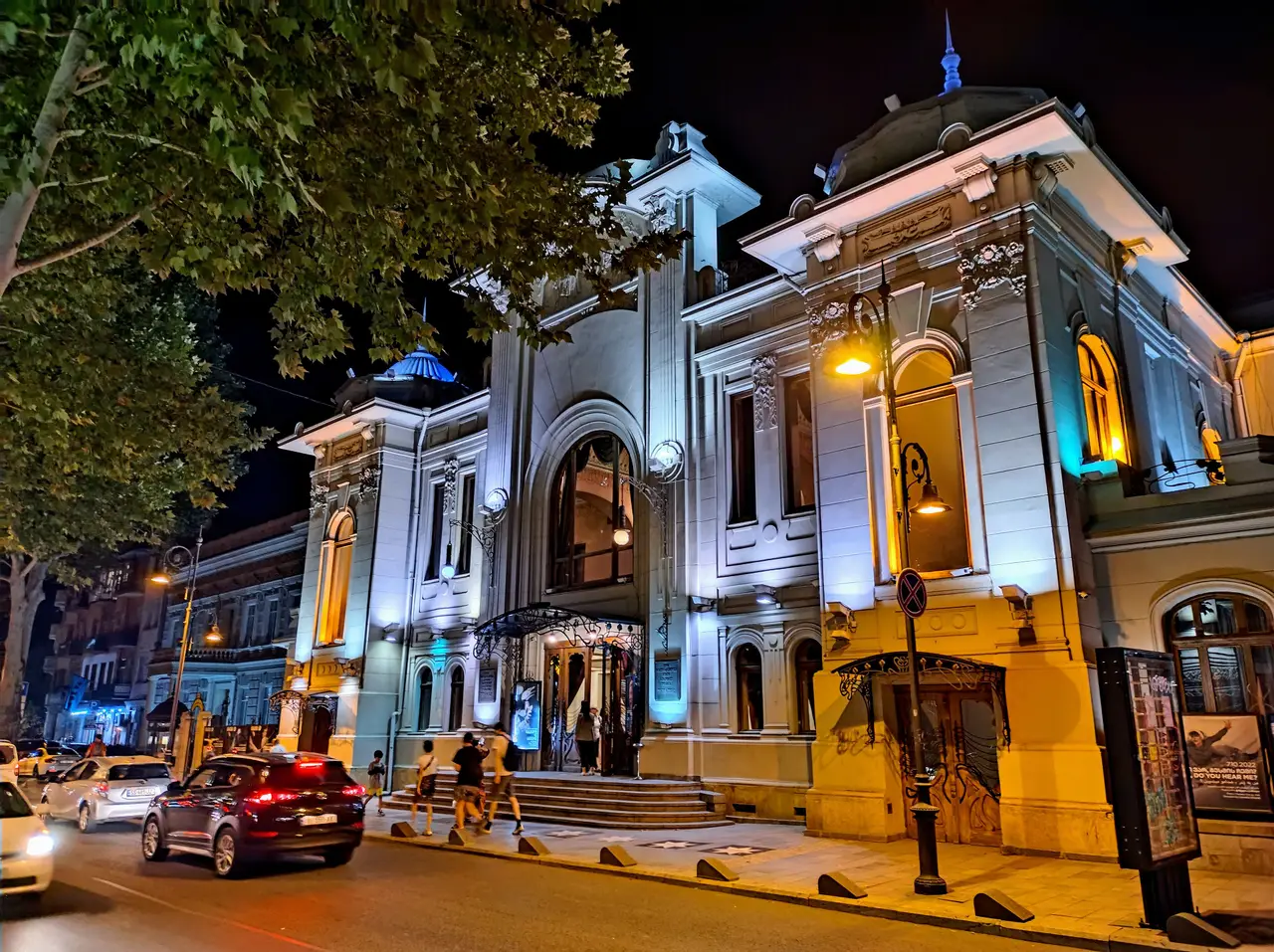 The white facade of the Marjanishvili Theatre in Tbilisi lit up at night, with a car passing on the street in front