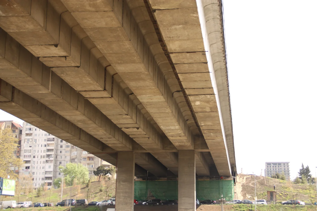 The concrete underside of the Vakhushti Bagrationi Bridge in Tbilisi, with apartment blocks and parked cars beyond the pillars