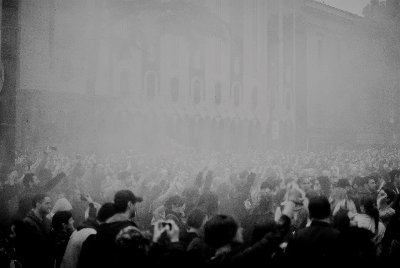 Black-and-white photo of a dense crowd with raised arms in smoke in front of the Parliament building in Tbilisi