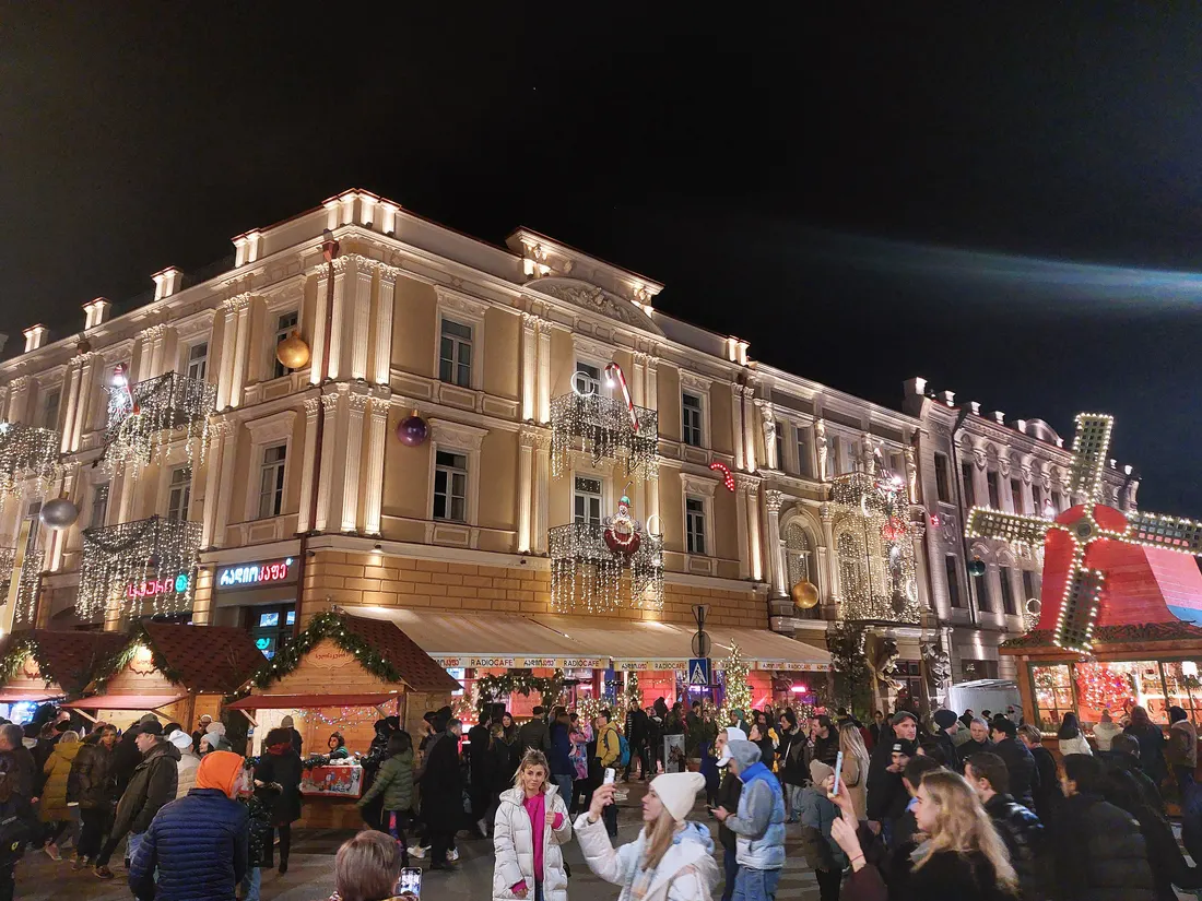 A lit-up 19th-century building on Atoneli Street in Tbilisi at night, hung with holiday lights, with market huts and a crowd below
