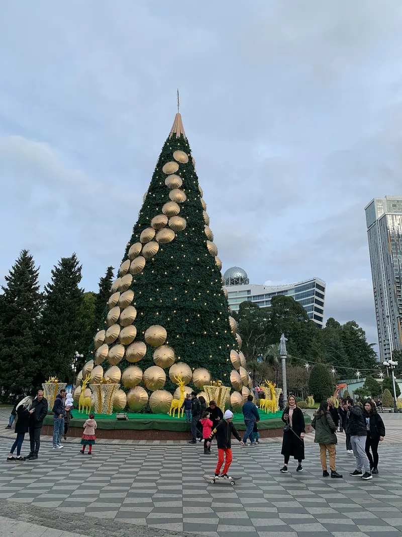 A tall New Year tree decorated with gold discs and lights on a paved square in Batumi, with people walking around it