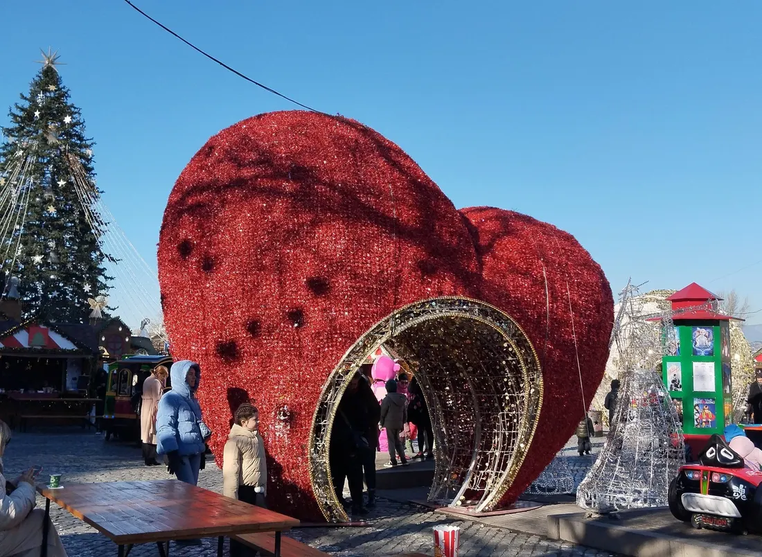 A giant red heart-shaped walk-through installation next to the main Christmas tree in Tbilisi's New Year Village, with families in winter coats