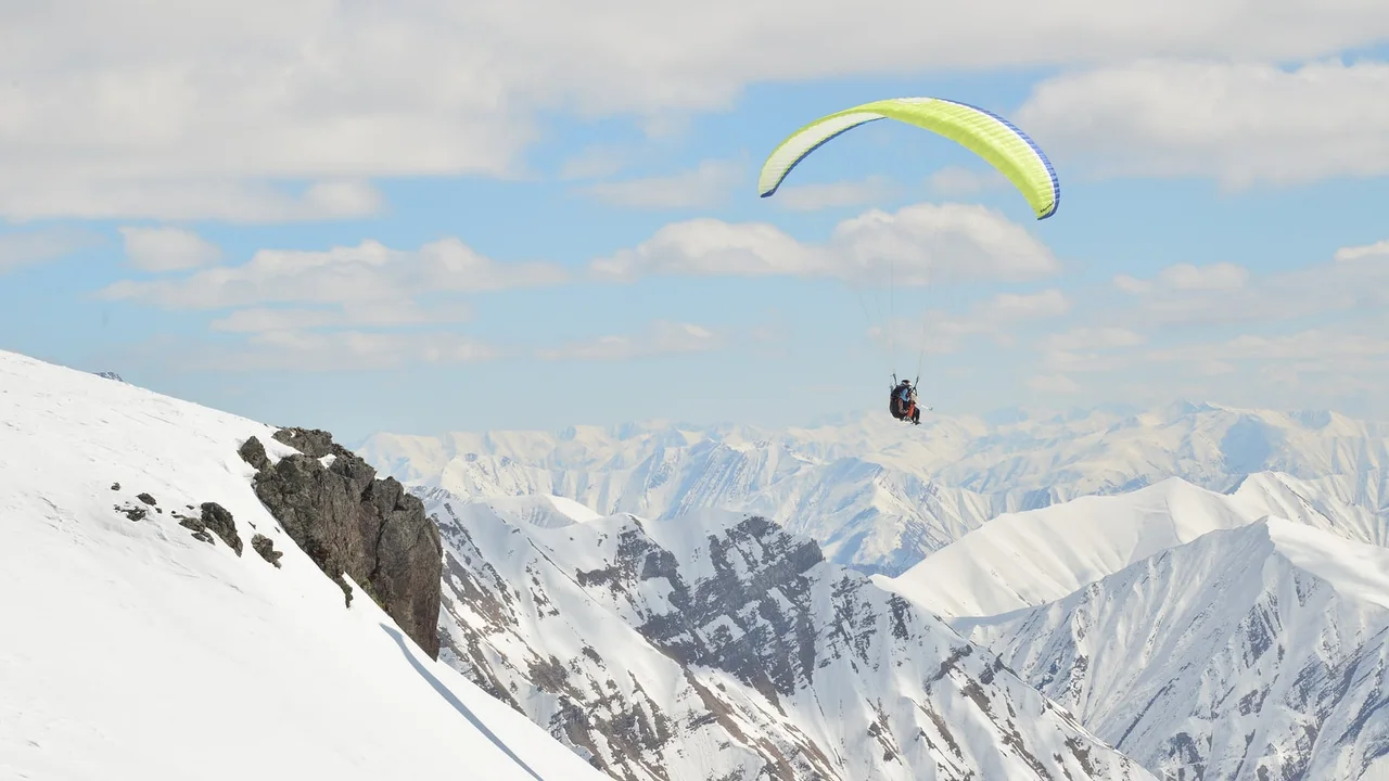A tandem paraglider with a yellow-green wing flying above snowy ridges at Gudauri