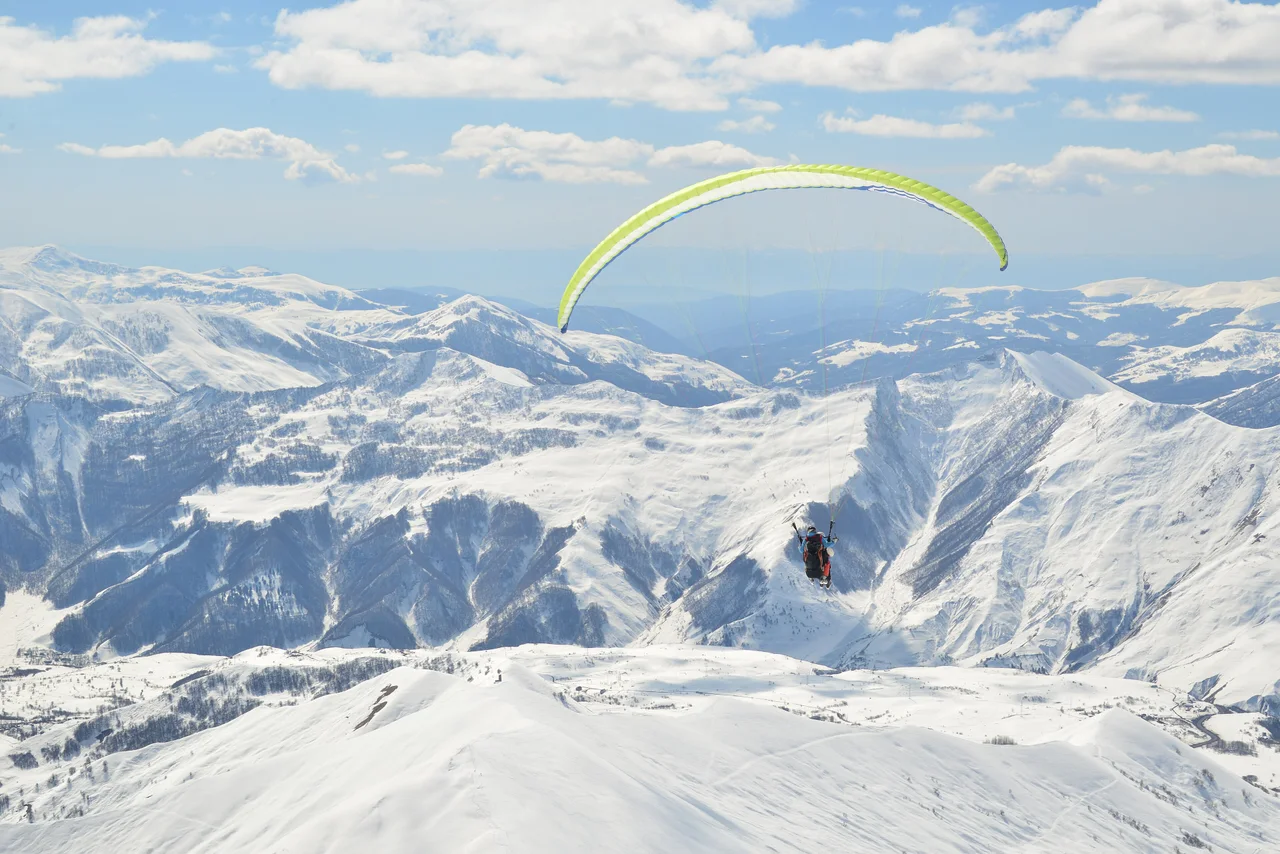 A tandem paraglider under a yellow-green wing above snowy Gudauri ridges