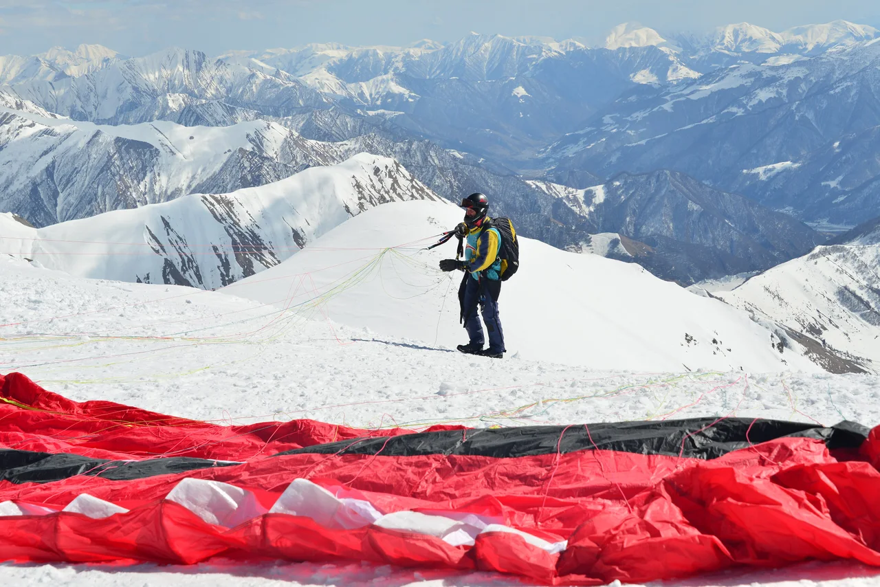 A pilot sorting lines beside a red wing laid out on the snow at a Gudauri launch