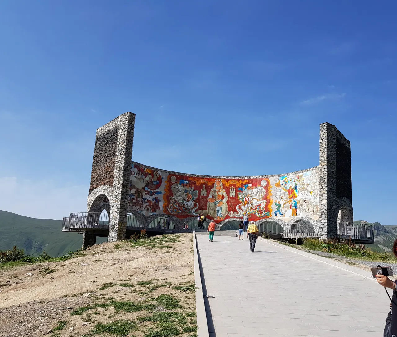 The Russia-Georgia Friendship Monument, a curved stone mosaic wall, with visitors walking up to it