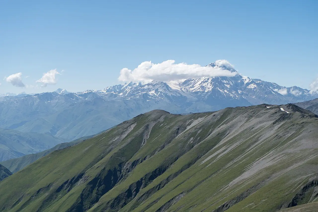 Kazbek seen from afar over a green mountain ridge, its summit wreathed in cloud
