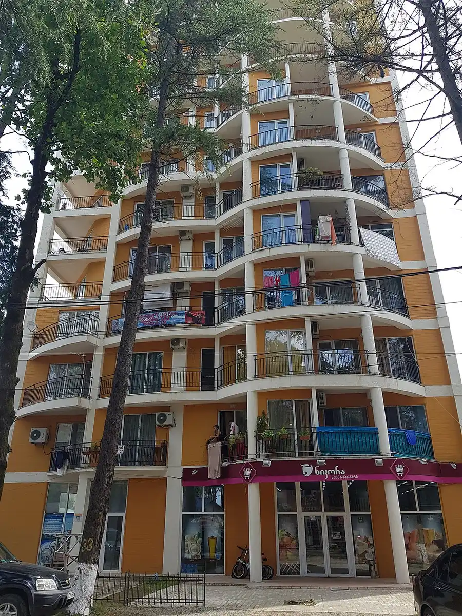 A tall orange guesthouse block in Ureki with balconies and a shop on the ground floor among pines